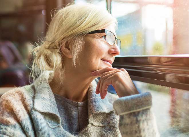 An older woman wearing glasses is looking out of a bus window